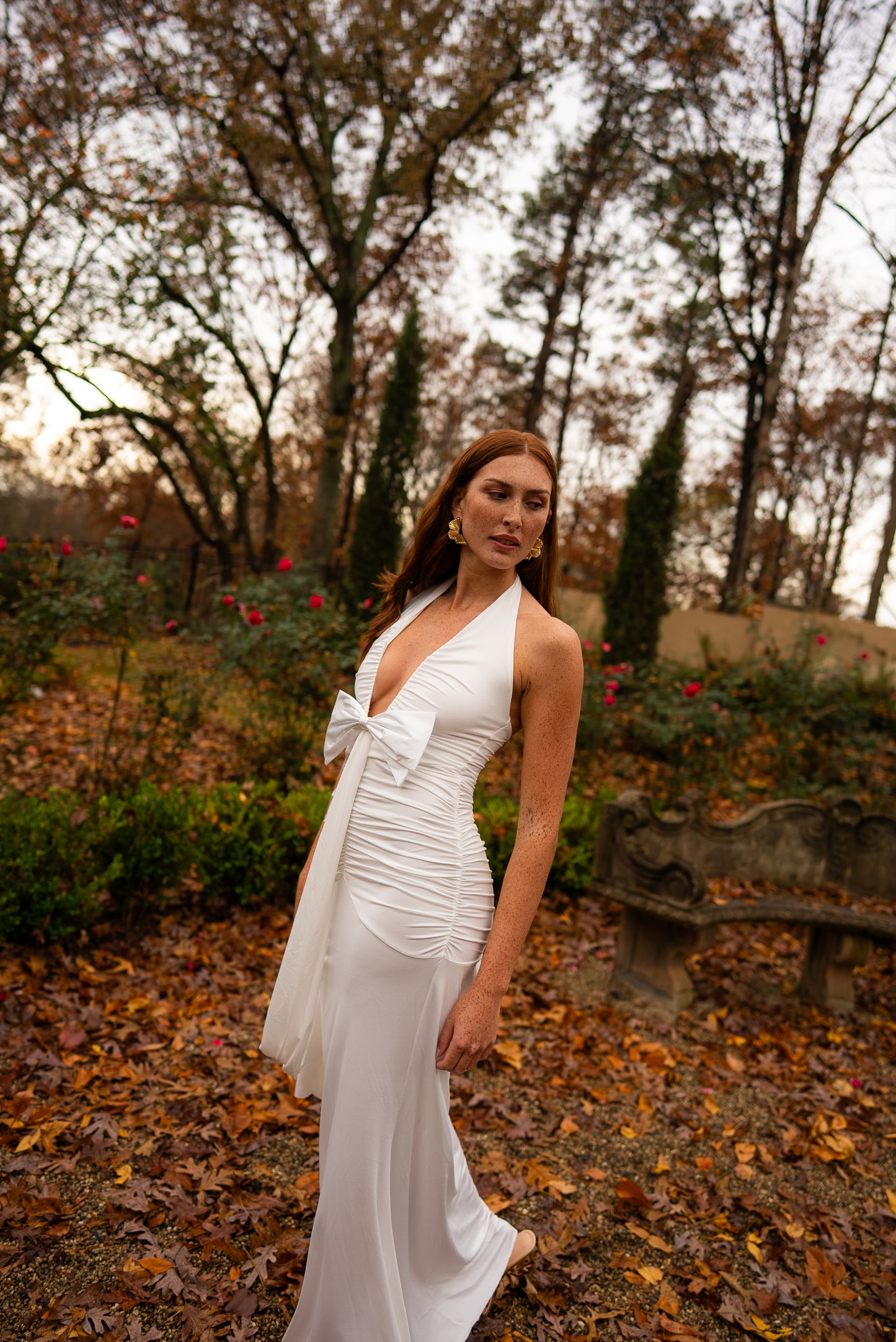 Woman in a white dress standing in a garden with trees and fallen leaves.