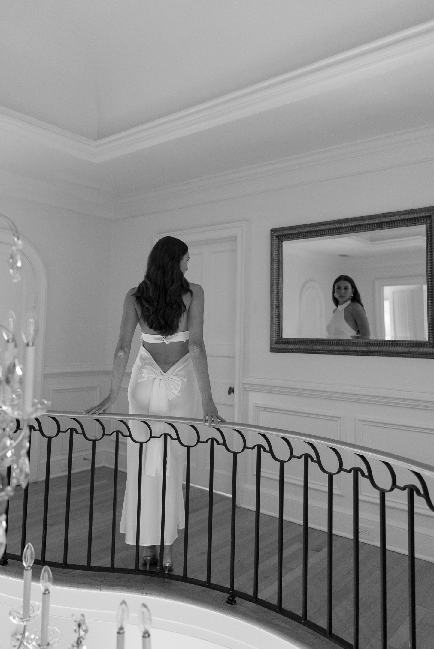 Woman in a white dress standing in a room with a mirror reflecting her image.