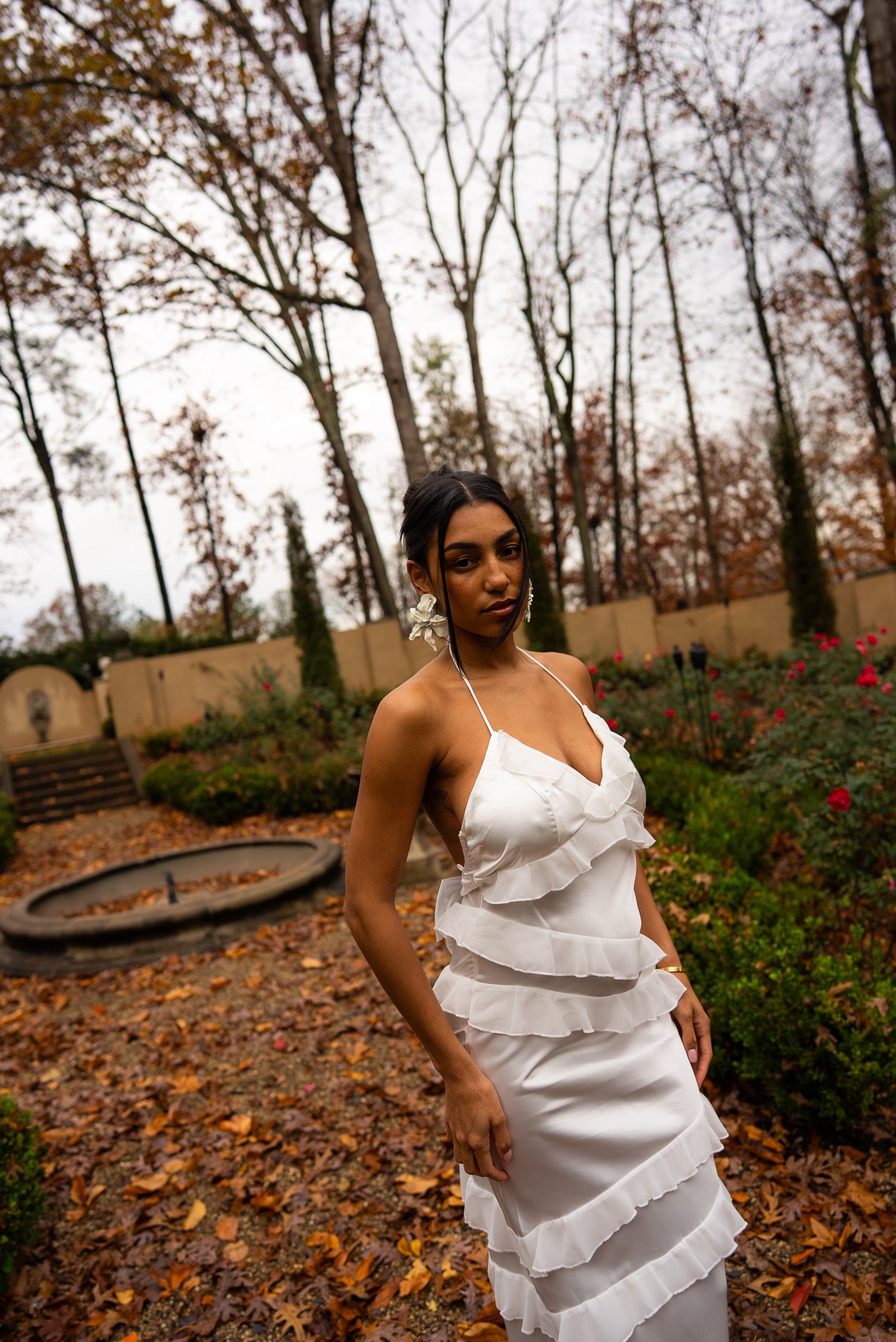 Woman in a white dress standing in a garden with bare trees and fallen leaves.