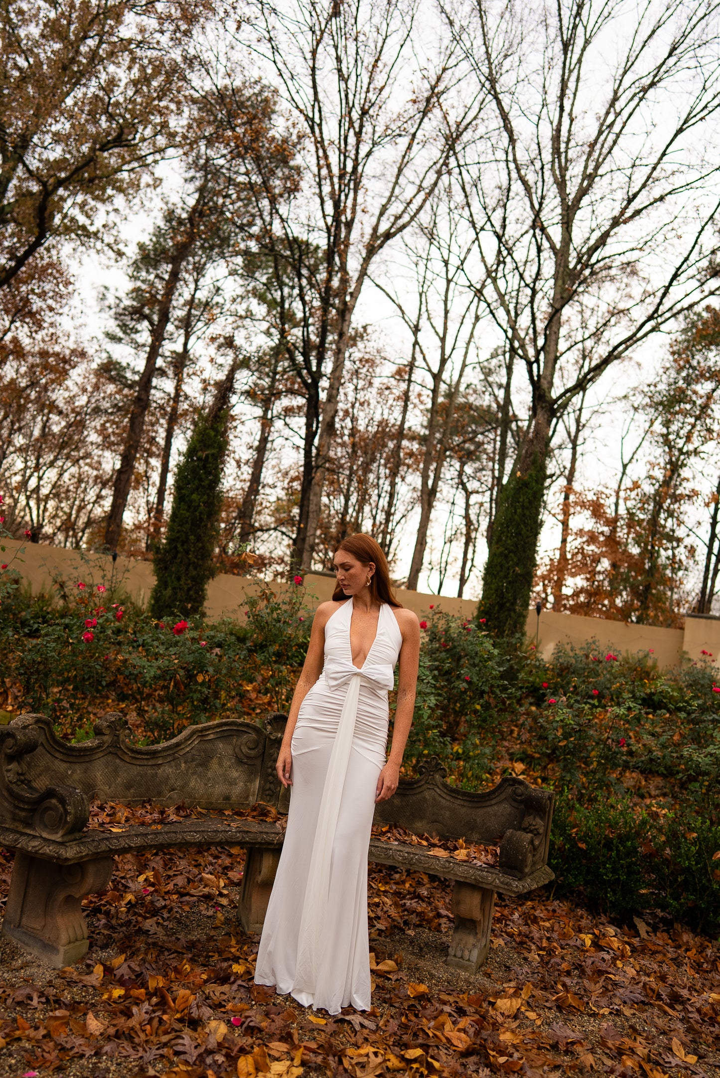 Woman in a white dress standing in an autumnal garden with bare trees and fallen leaves.