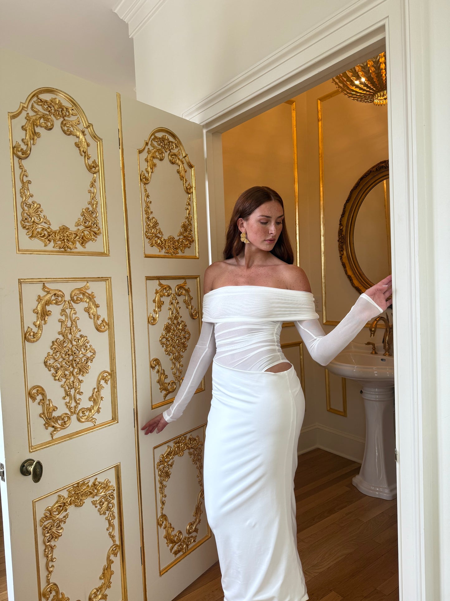 Woman in a white off-shoulder dress standing in a room with ornate gold decorations.