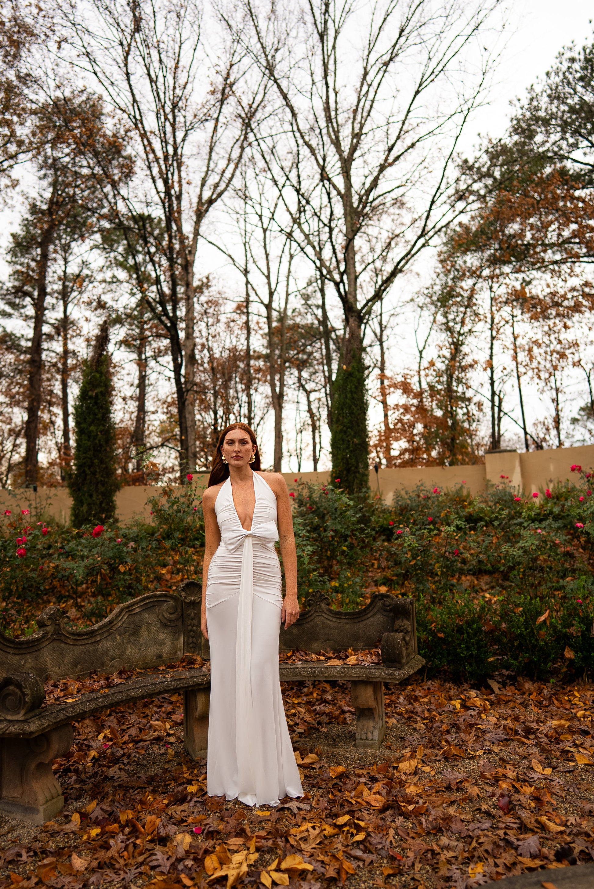 Woman in a white dress standing in an autumnal garden with bare trees and fallen leaves.